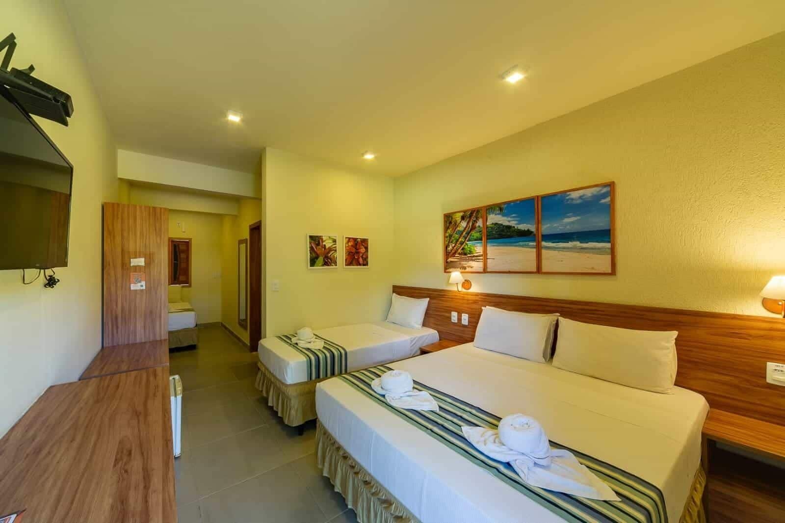 Quarto de hotel com duas camas, uma televisão e um quadro de uma praia.