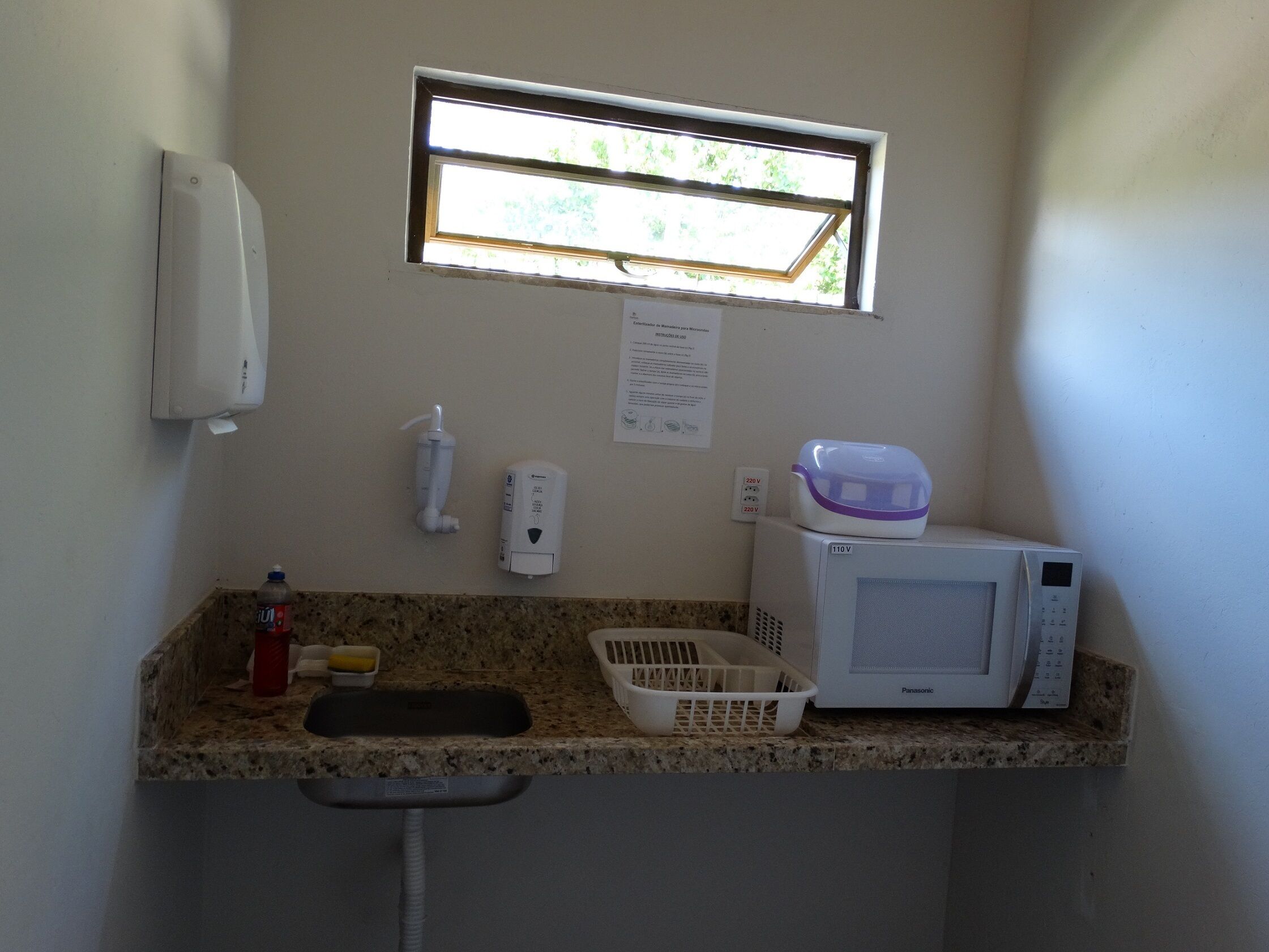 A kitchenette with a microwave, a small window, a hand sanitizer dispenser, a paper towel dispenser, and a water bottle.