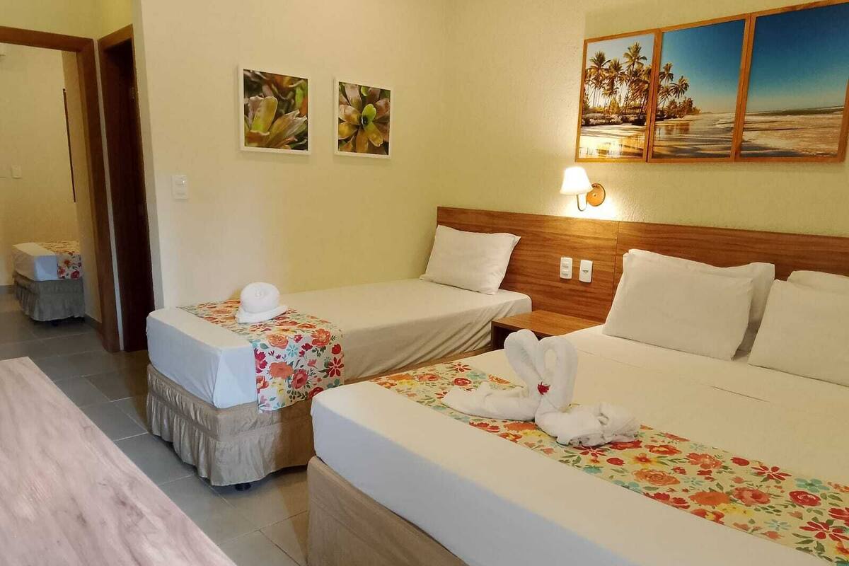 Ein Hotelzimmer mit zwei Einzelbetten, einem hölzernen Kopfteil, einem großen Bett mit floralem Überwurf und einem Bild einer Strandlandschaft an der Wand.