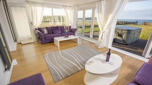 Amplia sala de estar con un ventanal, un sofá morado, una mesa de centro blanca y una mesa redonda con una botella y vasos.