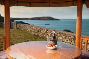 A round wooden table with a bottle of wine, two glasses, and a bowl of strawberries. - Seaside Luxury Villa Cottage with Hot Tub  and stunning uninterrupted sea views! (CEMAES BAY)