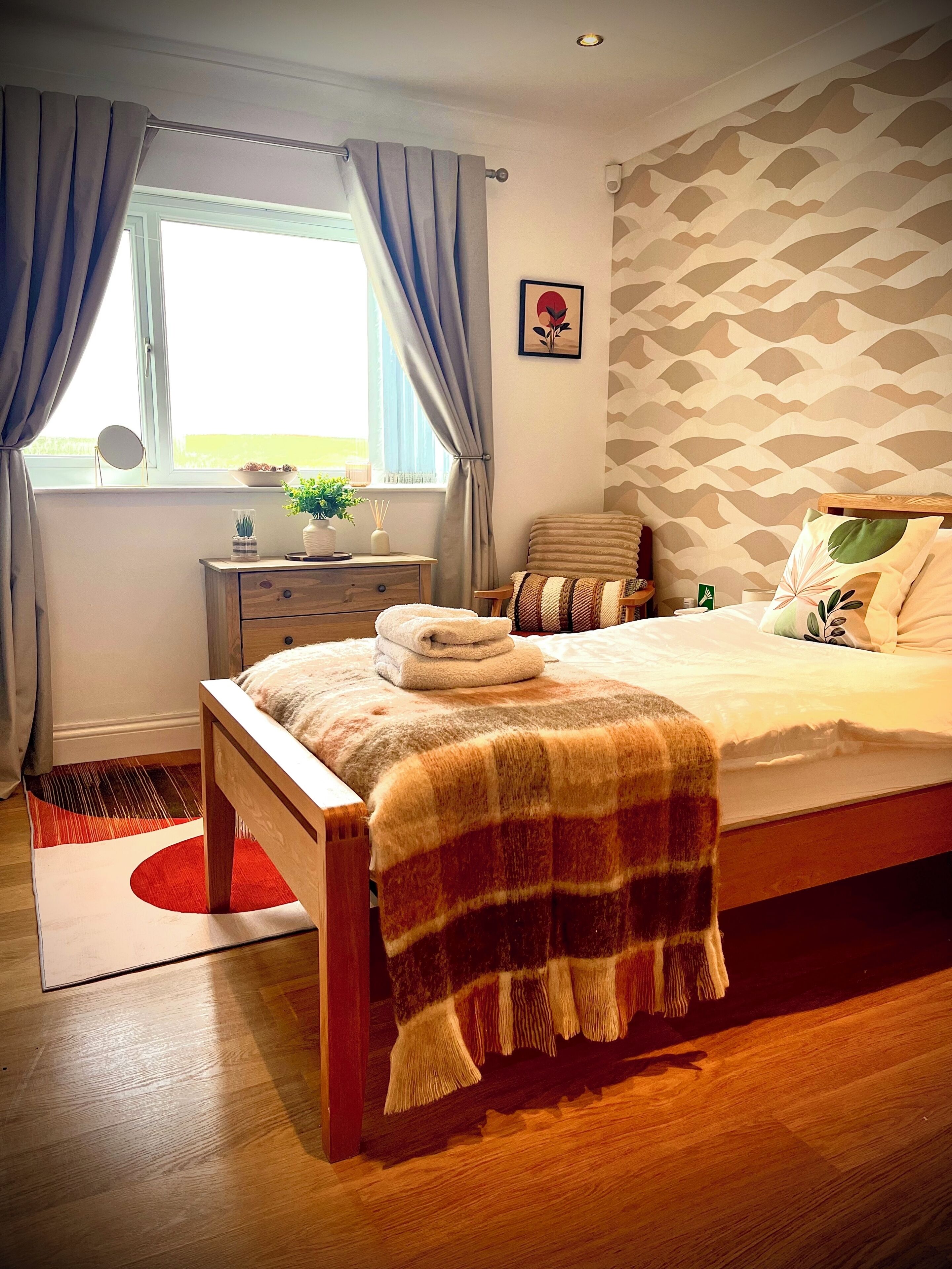 A bedroom with a bed, a bedside table, a window with curtains, a wall with a geometric pattern, and a small plant on the window sill.