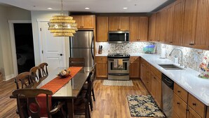 Une cuisine moderne avec des meubles en bois, des appareils électroménagers en acier inoxydable et un coin repas comprenant une table en verre et des chaises en bois.