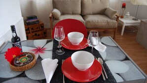 Une table à manger est dressée avec des assiettes rouges, des bols blancs et des verres à vin. Une bouteille de vin de Madère et un panier de victuailles sont posés sur le côté gauche.