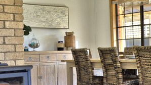Un coin repas avec une table en bois et quatre chaises en osier, un mur en briques, une lampe décorative et une fenêtre avec des stores.