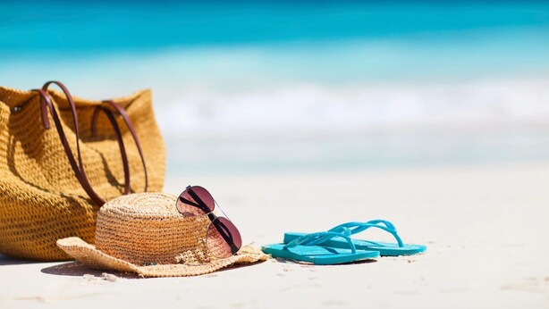 Un chapeau de paille, des lunettes de soleil et des tongs bleues sur une plage de sable, avec un océan bleu et limpide en arrière-plan.