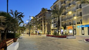 Ein gepflasterter Fußweg mit Palmen, Bänken und Gebäuden mit Balkonen und Beschilderung.