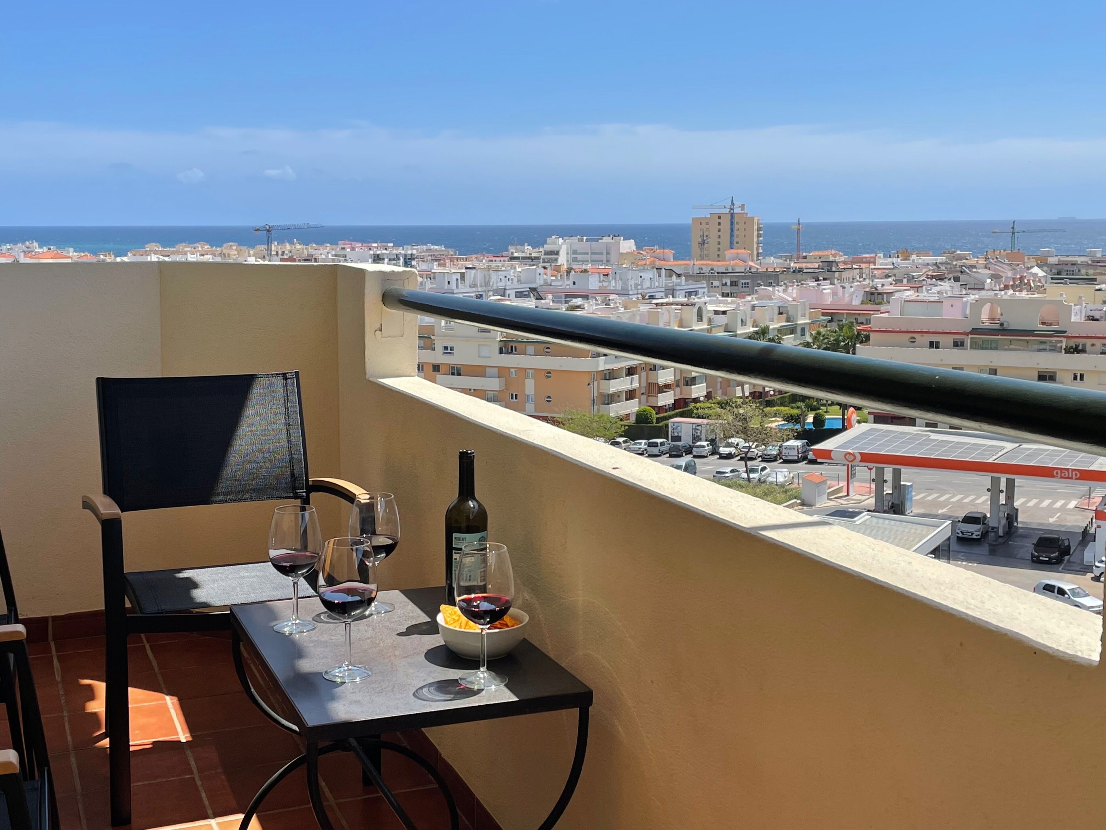 Un balcon avec une table dressée pour deux personnes, une bouteille de vin et un bol de collations. Le balcon offre une vue sur le paysage urbain, avec ses bâtiments et son ciel dégagé.