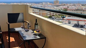 Ein Balkon mit Tisch für zwei Personen, einer Weinflasche und einer Schale mit Snacks. Der Balkon bietet Blick auf eine Stadtansicht mit Gebäuden und wolkenlosem Himmel.