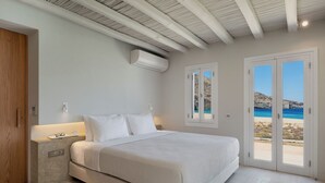 A bedroom with a large bed, a nightstand, a wooden door, and a view of the beach through a glass door.