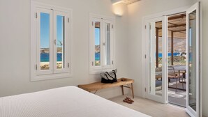 A bedroom with a bed, a wooden bench, a pair of sandals, and a view of the sea through a sliding glass door.