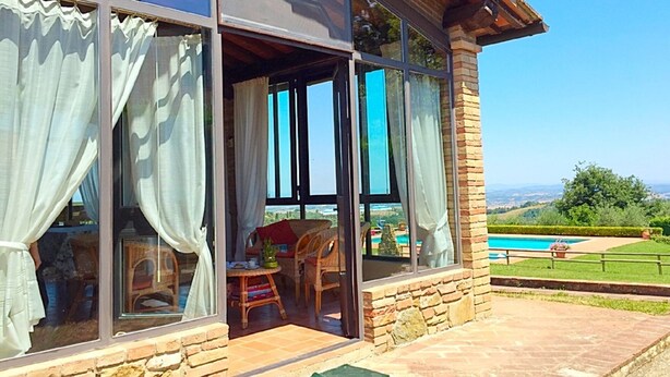 A modern house with large glass windows, a patio area with a table and chairs, and a view of a swimming pool and landscape.