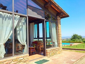 A modern house with large glass windows, a patio area with a table and chairs, and a view of a swimming pool and landscape.