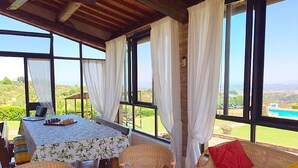 A covered outdoor seating area with wicker furniture, a table with a chessboard, and large windows offering a view of a pool and landscape.