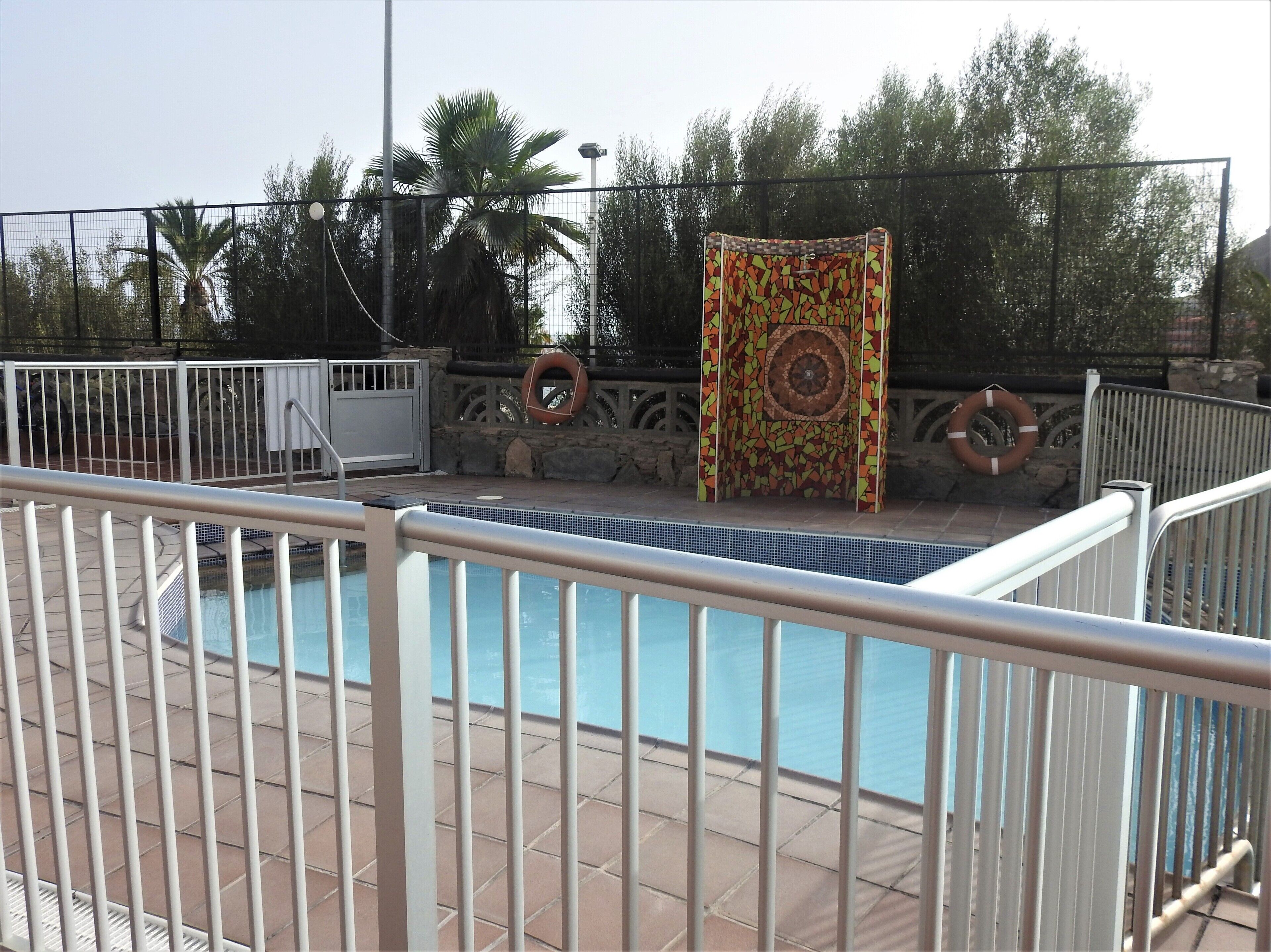 Una piscina con plataforma empedrada, rodeada por una valla metálica. Se observan chalecos salvavidas sujetos a la valla, y una puerta con diseño es visible al fondo.