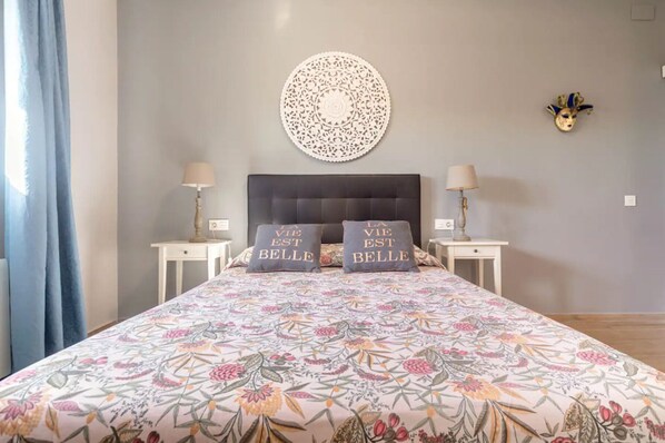 A bedroom with a floral bedspread, two bedside tables with lamps, a round wall decoration, and two pillows with the text "LA VIE EST BELLE".