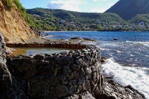 A coastal landscape with a rocky shoreline, a clear blue sea, and a backdrop of hills.