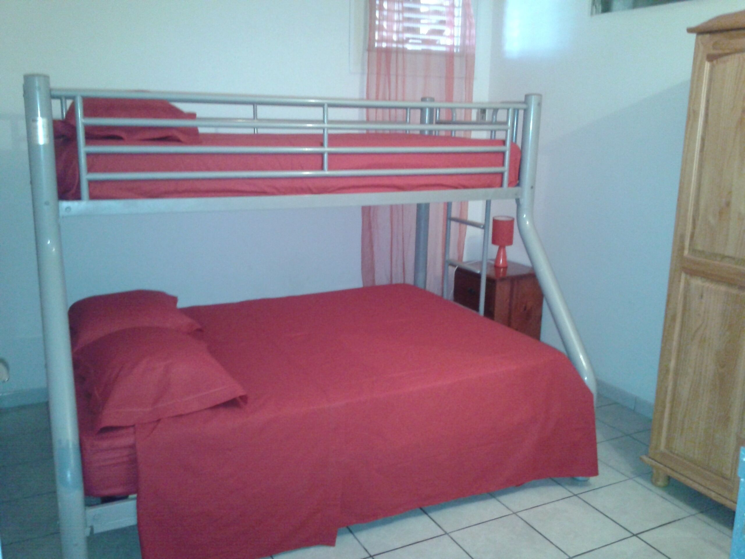A bunk bed with red bedding in a room with a wooden cabinet and a window with curtains.