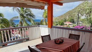 A covered outdoor dining area with a table and chairs, a view of the ocean, and a hilly landscape in the background.