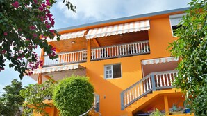 A two-story building with a bright orange facade, a wooden balcony, and a small garden area with a tree and shrubs.