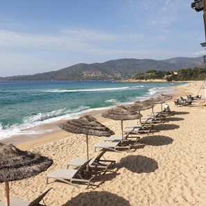 A beach with sun loungers and thatched umbrellas, a clear view of the sea, and a distant mountain range.