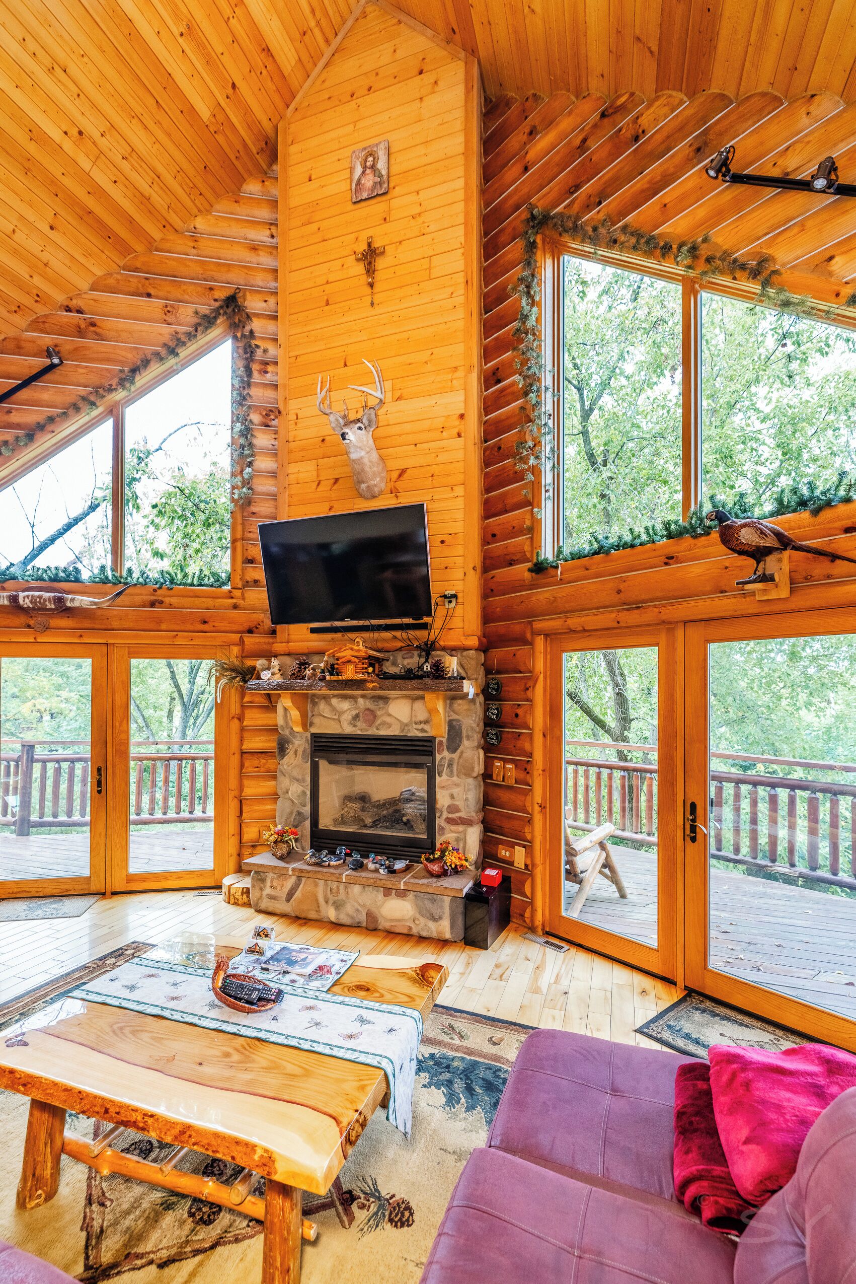 L’intérieur d’une cabane en rondins, avec une cheminée en pierre, un téléviseur fixé au mur et une grande fenêtre donnant sur une terrasse.