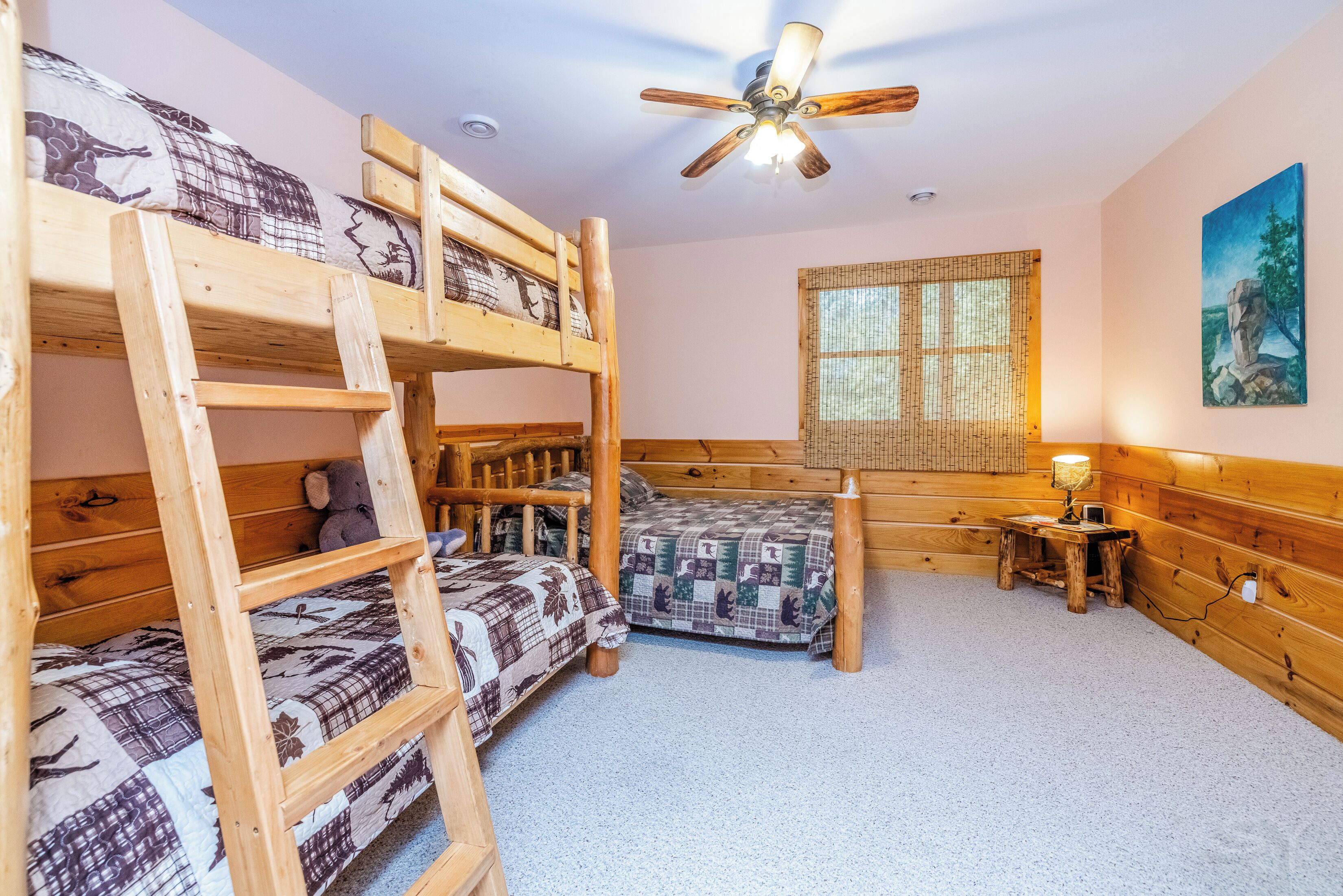A bunk bed room with a ceiling fan, a window with bamboo blinds, and a small table with a lamp.