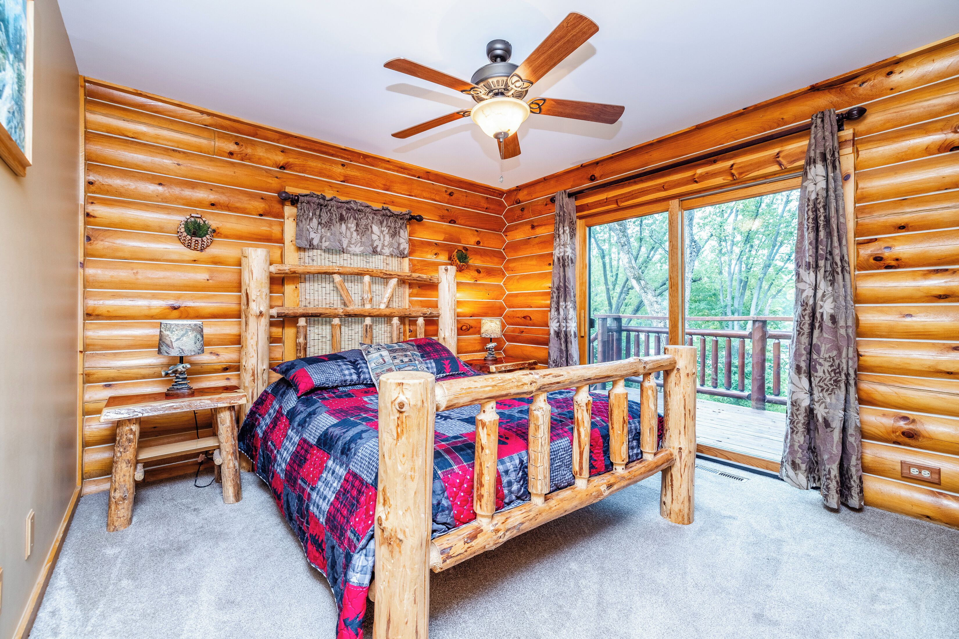 A log cabin bedroom with a bed, a ceiling fan, a window with curtains, and a view of trees.