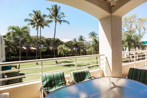 A balcony with a glass table and striped chairs, overlooking a lawn with palm trees and a clear blue sky.