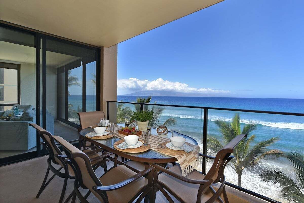 Uma varanda com mesa posta para refeições, com vista para uma praia com ondas e céu claro.