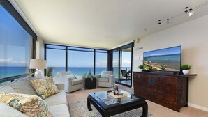 Ein modernes Wohnzimmer mit großem Fenster mit Blick auf den Ozean, einer gemütlichen Sofagarnitur, einem Holztisch und einem Flachbildfernseher auf einer Holzkommode.