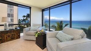 Ein modernes Wohnzimmer mit großem Fenster mit Blick auf Strand und Meer. Es gibt ein beiges Sofa, einen kleinen Beistelltisch und einen dekorativen Schrank.