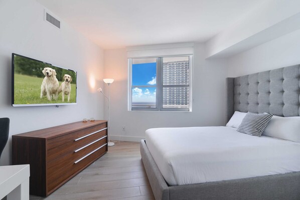 A modern bedroom with a large bed, a wooden dresser, a flat-screen TV, and a window with a view of a cityscape.