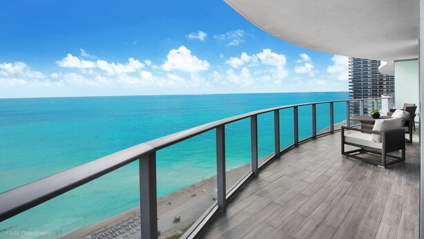 Ein Balkon mit Blick auf den Ozean, eine Sitzecke mit einer Couch und Stühlen, sowie ein modernes Gebäude im Hintergrund.