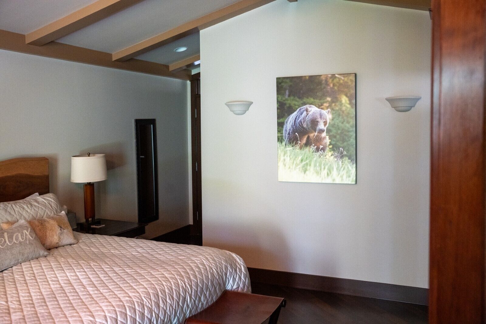 A bedroom with a bed, a bedside table with a lamp, a flat-screen TV, and a wall-mounted picture of a bear in a forest.