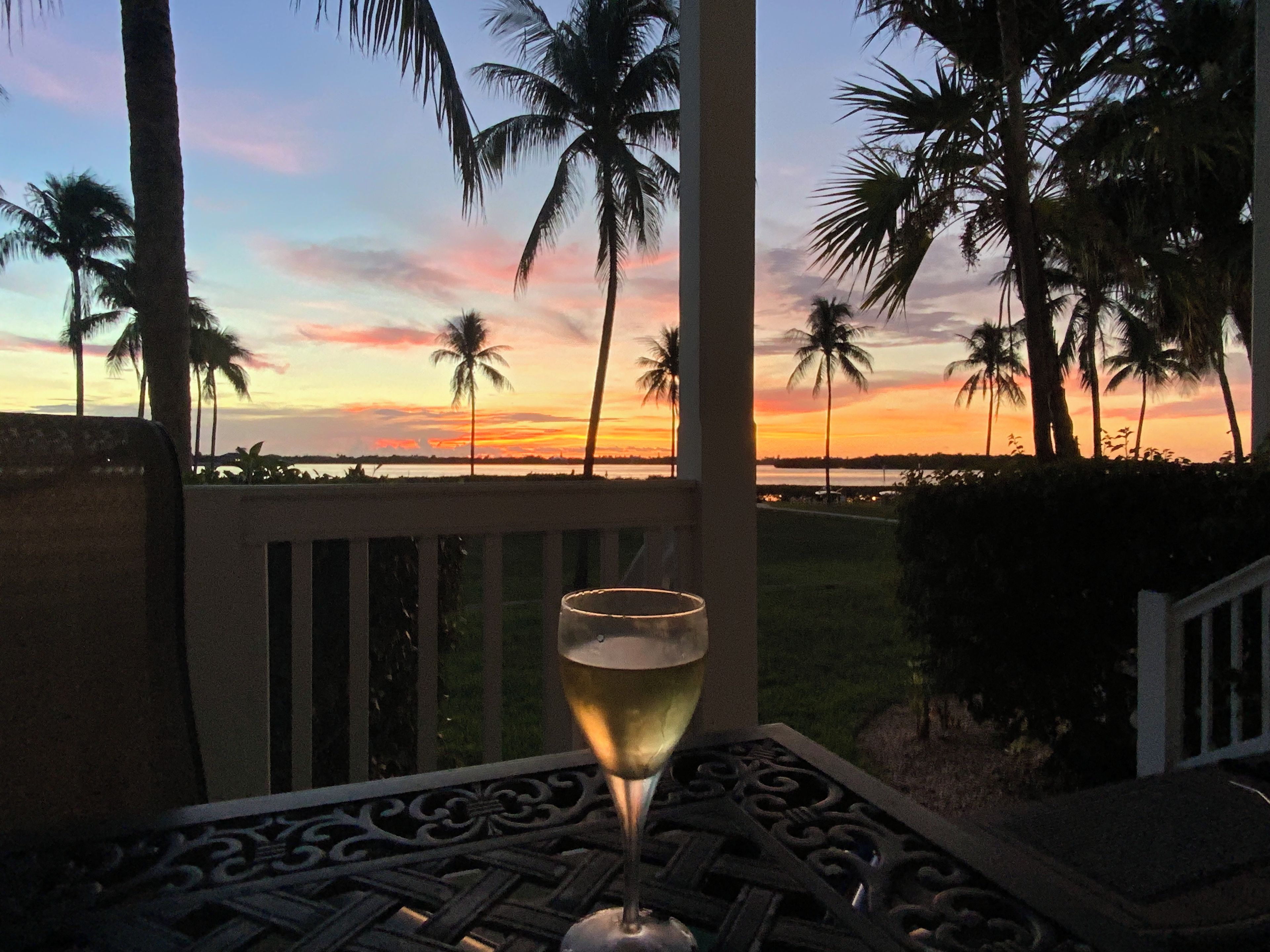 Une table avec un verre de vin blanc, donnant sur un coucher de soleil avec des palmiers en arrière-plan.