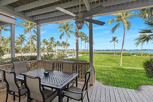 A covered outdoor seating area with a table and chairs, overlooking a grassy lawn and palm trees, with a view of the ocean and buildings in the distance.