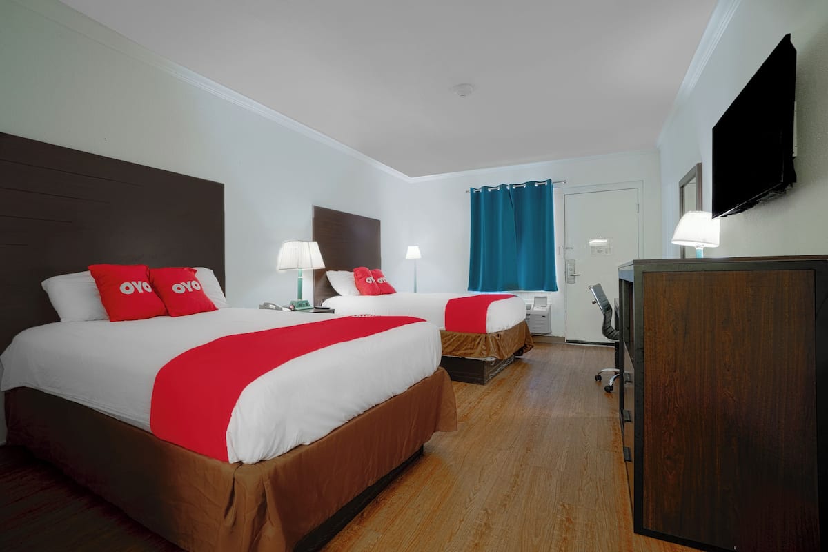 Ein Hotelzimmer mit zwei Betten, jeweils mit roter und weißer Bettwäsche, Holzboden, Schreibtisch mit Stuhl, Wand-Fernseher und einem Fenster mit blauen Vorhängen.