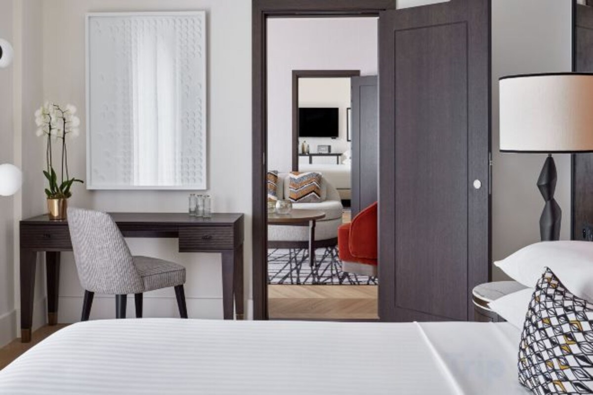 A hotel room with a bed, a desk, a chair, a lamp, and a view of a dining area.