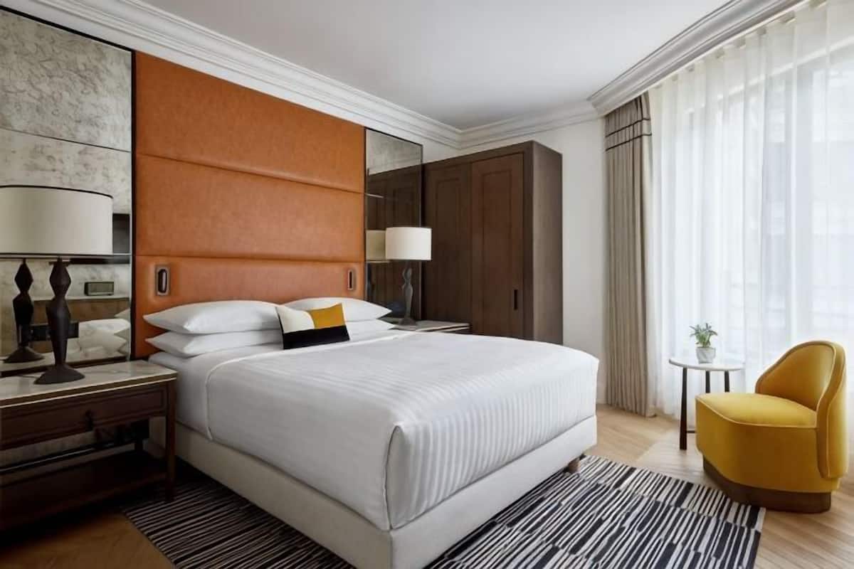 Una habitación de hotel con una cama grande, una mesita de noche, una silla, una pequeña mesa con una planta y un ventanal con cortinas.