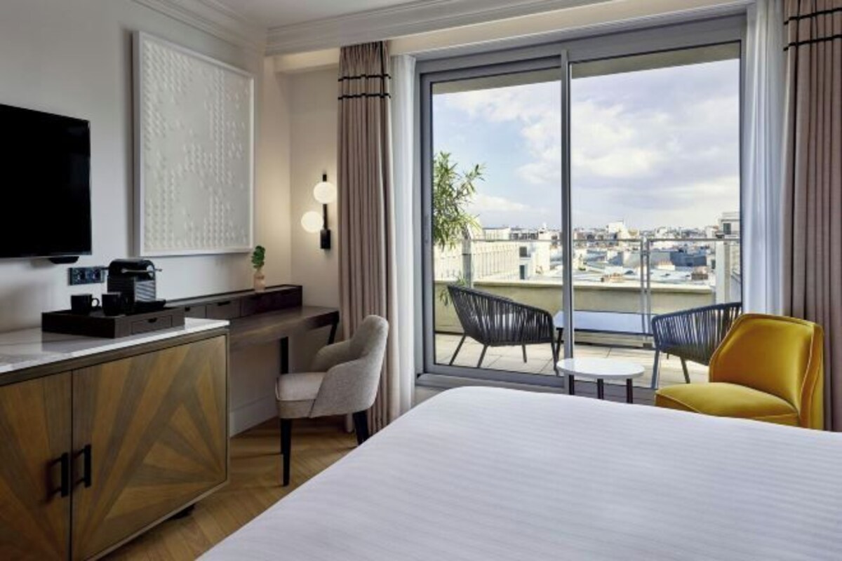 Een hotelkamer met een bed, een bureau met een flatscreen televisie, een stoel, een klein tafeltje en een balkon met stoelen.