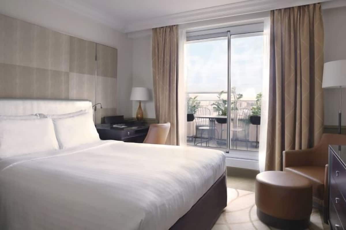 Habitación de hotel con una cama grande, un escritorio, una silla, un pequeño reposapiés y un balcón con vistas.