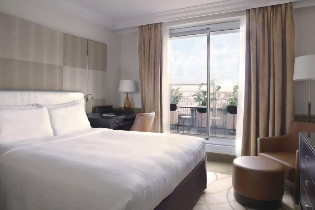Ein Hotelzimmer mit einem großen Bett, einem Schreibtisch, einem Sessel, einem kleinen Hocker und einem Balkon mit Blick.