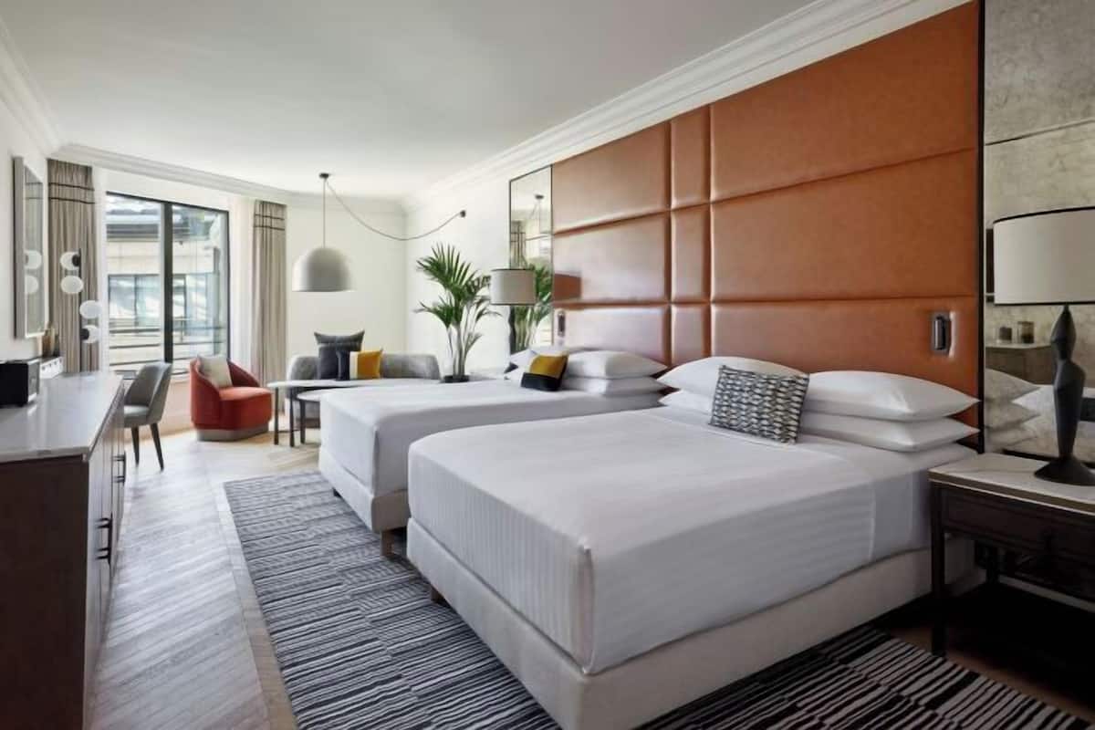 Habitación de hotel con dos camas, un escritorio, una silla, un espejo grande y vistas a la ciudad.