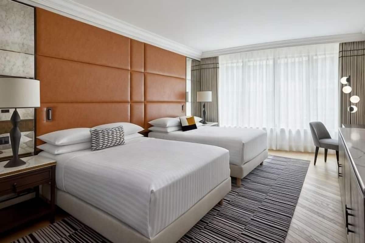 Una habitación de hotel con dos camas, un escritorio, una silla y un ventanal con cortinas.