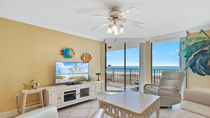 A living room with a TV, white furniture, a ceiling fan, and a view of the beach.