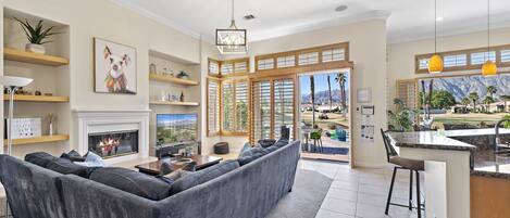 A modern living room with a fireplace, a large sofa, and a view of a mountainous landscape through the windows.