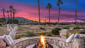A stone fire pit with a fire, surrounded by seating with cushions, palm trees, and a golf course in the background. - 3 Bed Permit#227096 PGA West, relaxing heated pool home with views! (La Quinta)