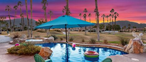Ein Pool mit Liegestühlen und einem blauen Sonnenschirm, umgeben von Palmen und einer Bergkulisse.