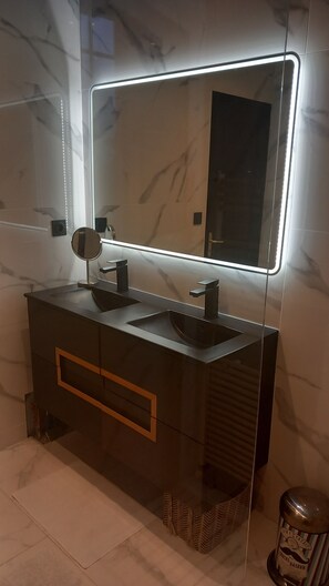 Een moderne badkamer met een donker badkamermeubel, twee wastafels, een grote spiegel met LED-verlichting en een prullenbak.